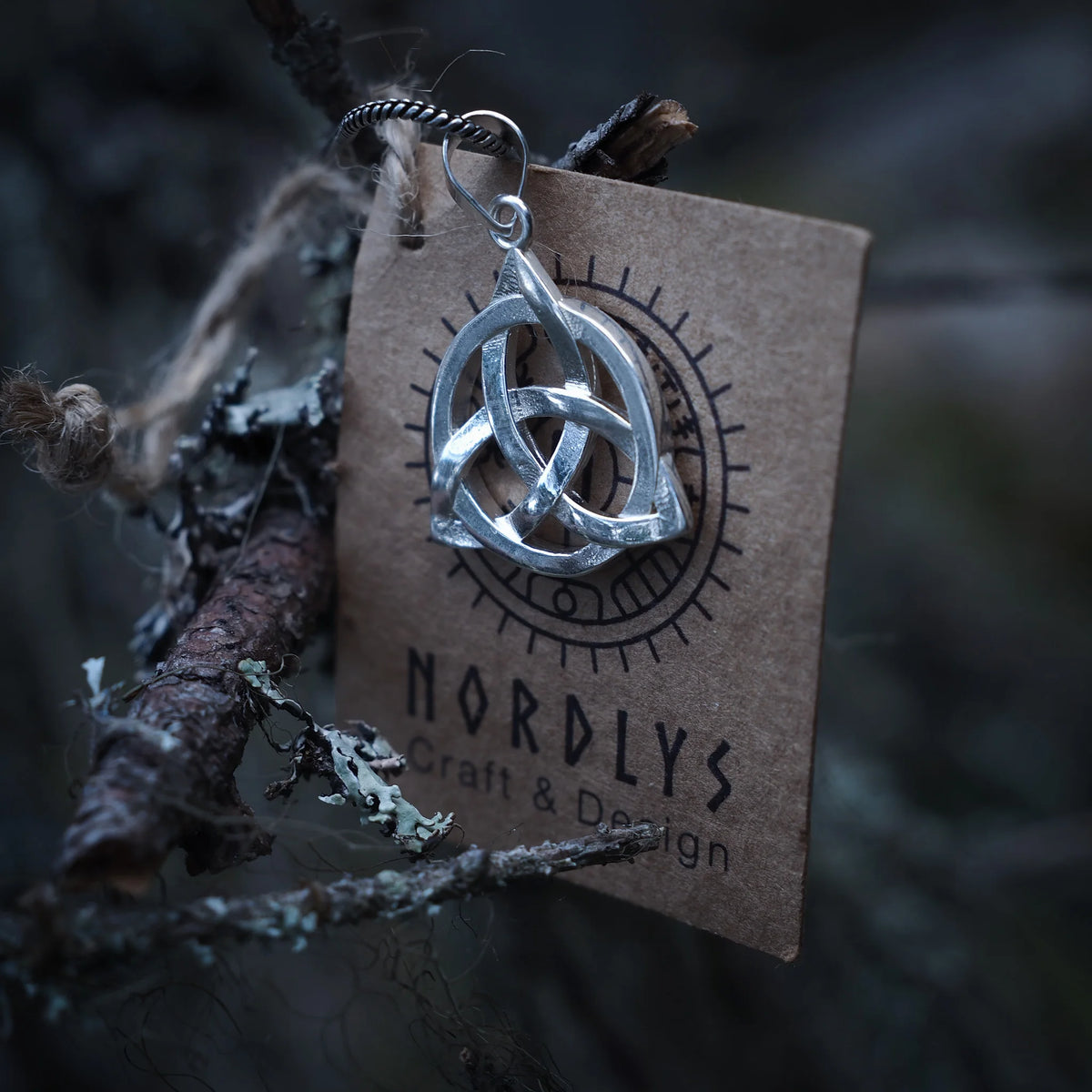 En silverbelagd evighetsknut-hängsmycke med trekantigt vävmönster, upphängd från en metallslinga framför en NORDLYS-märkt kraftpappersask med sollogga, omgiven av grenar och mossa