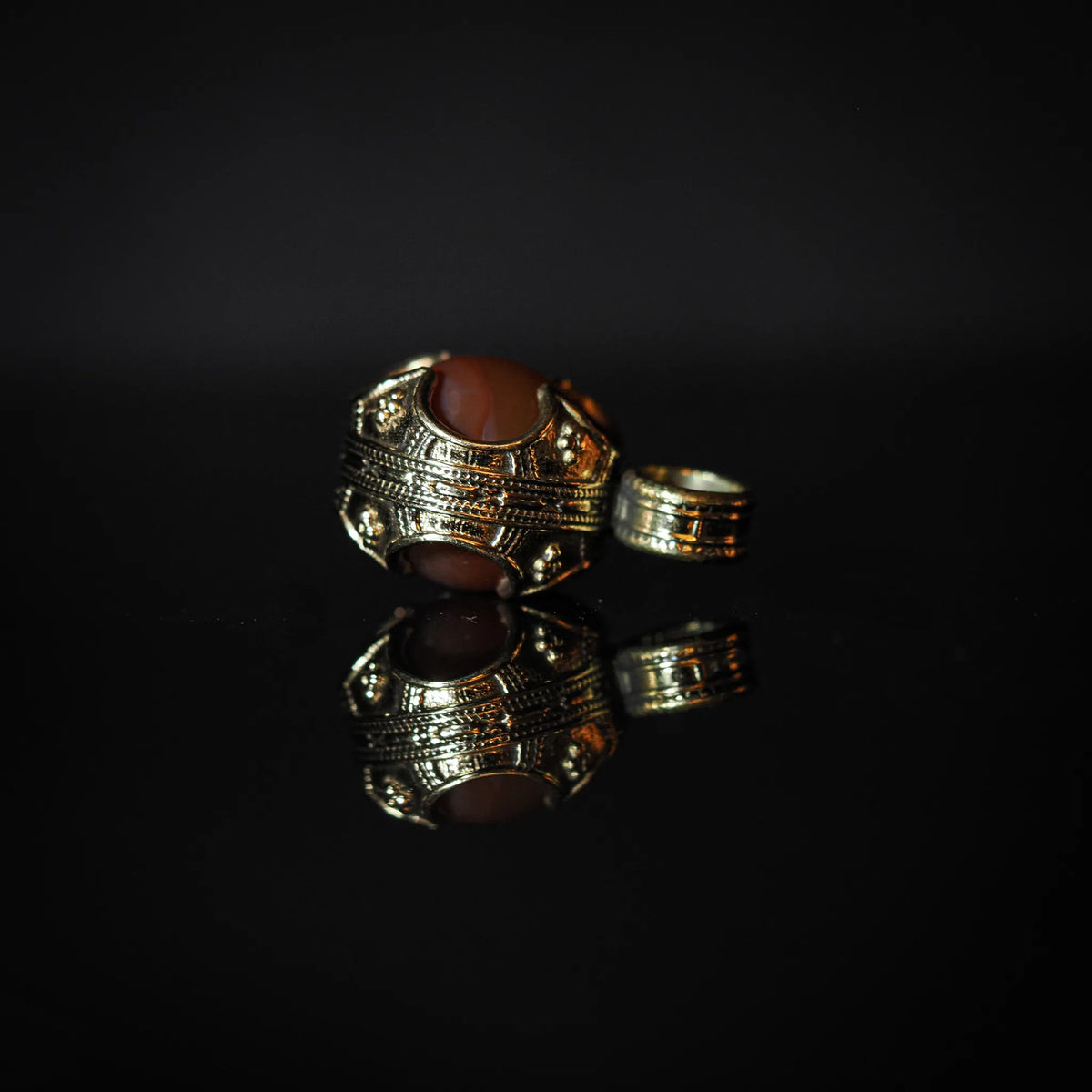 Närbild av ornerad silverpläterad vikingahänge med detaljerade streckmönster och glansig rödagat-sten i profil. Liten sling-ring syns i bakgrunden.