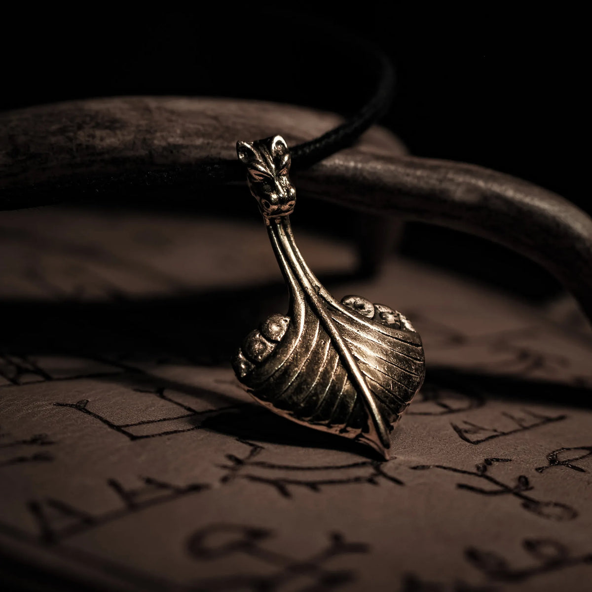 Bronshänge i form av vikingaskepp med drakehuvud vid förskepp. Hängande från svart lädersnöre. Detaljerad metallarbete med varm glans.