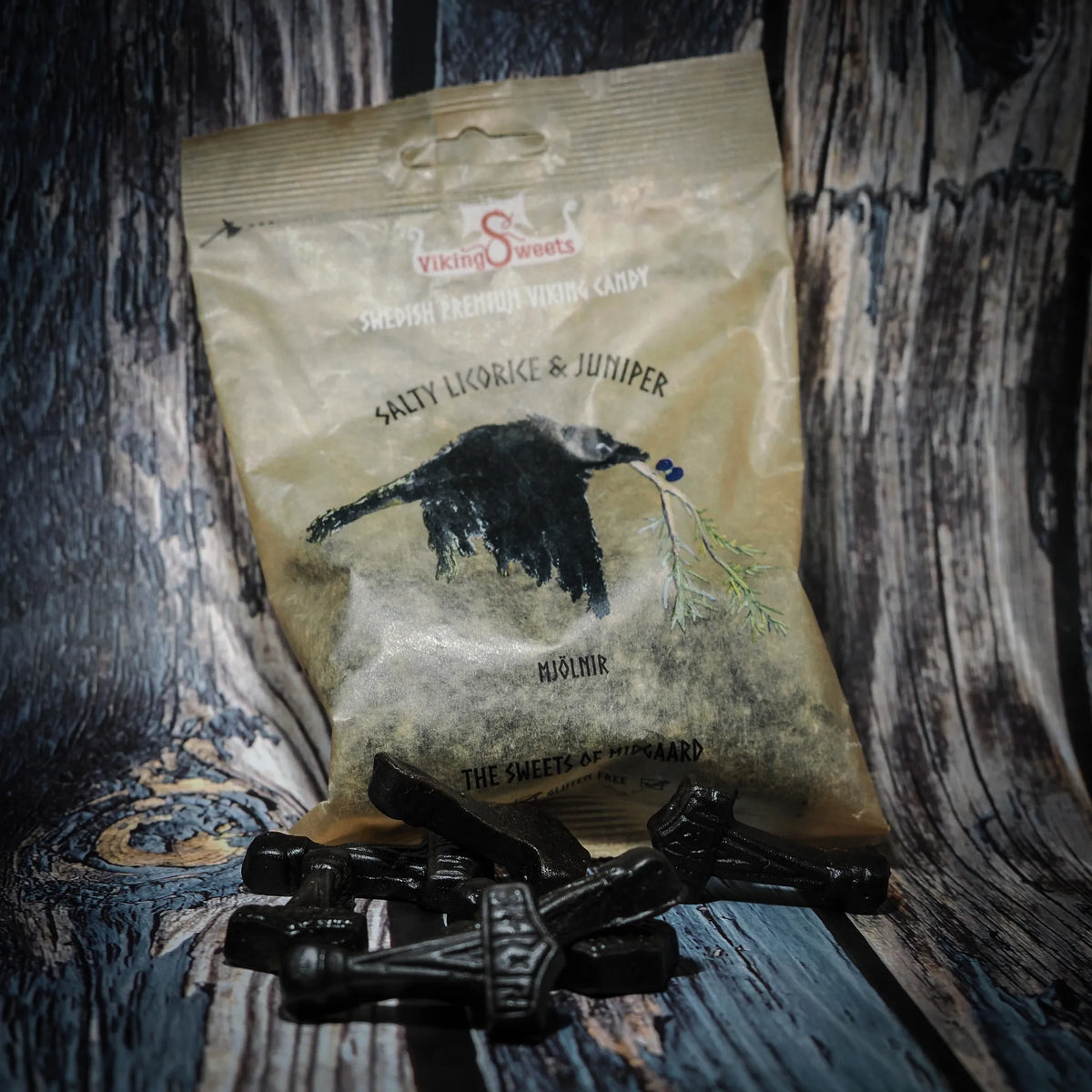Svarta Mjölnir-formade godisbitar framför en pastellgul förpackning med en svart råkorre och grön enriskvista. Vikinggodis i saltlakrits- och enrismaK presenterad på trä.