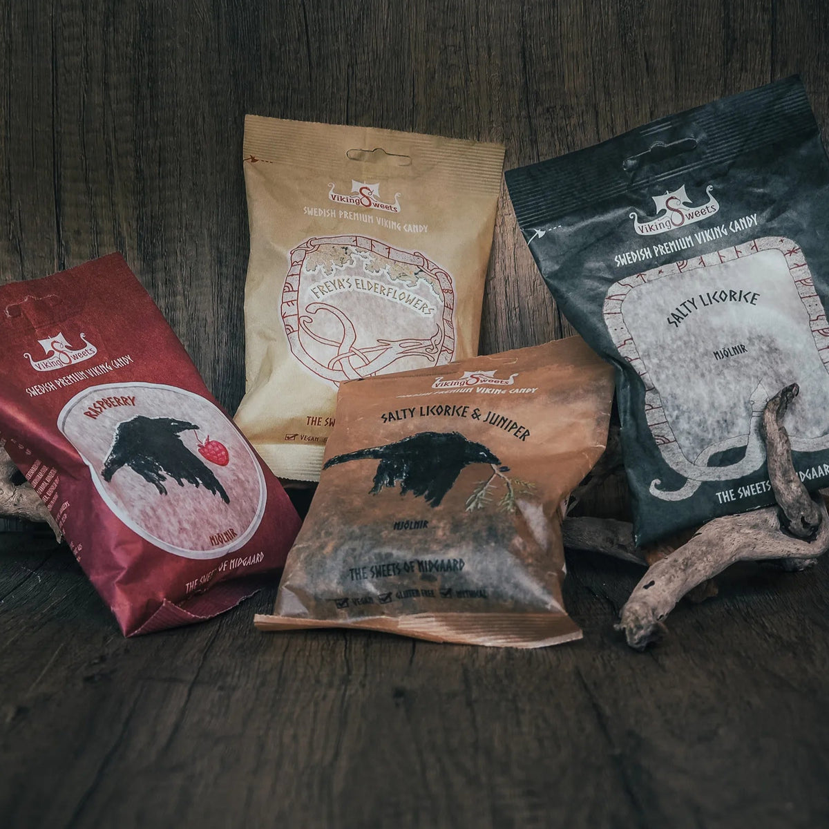 Fyra paket med vikinggodis i form av Mjölnir i olika smaker: hallon (röd), fläder (beige), saltlakrits med enbär (brun) och saltlakrits (marinblå). Paketen visar vikinginspirerad design med korp- och runmotiv på mörk träbakgrund.