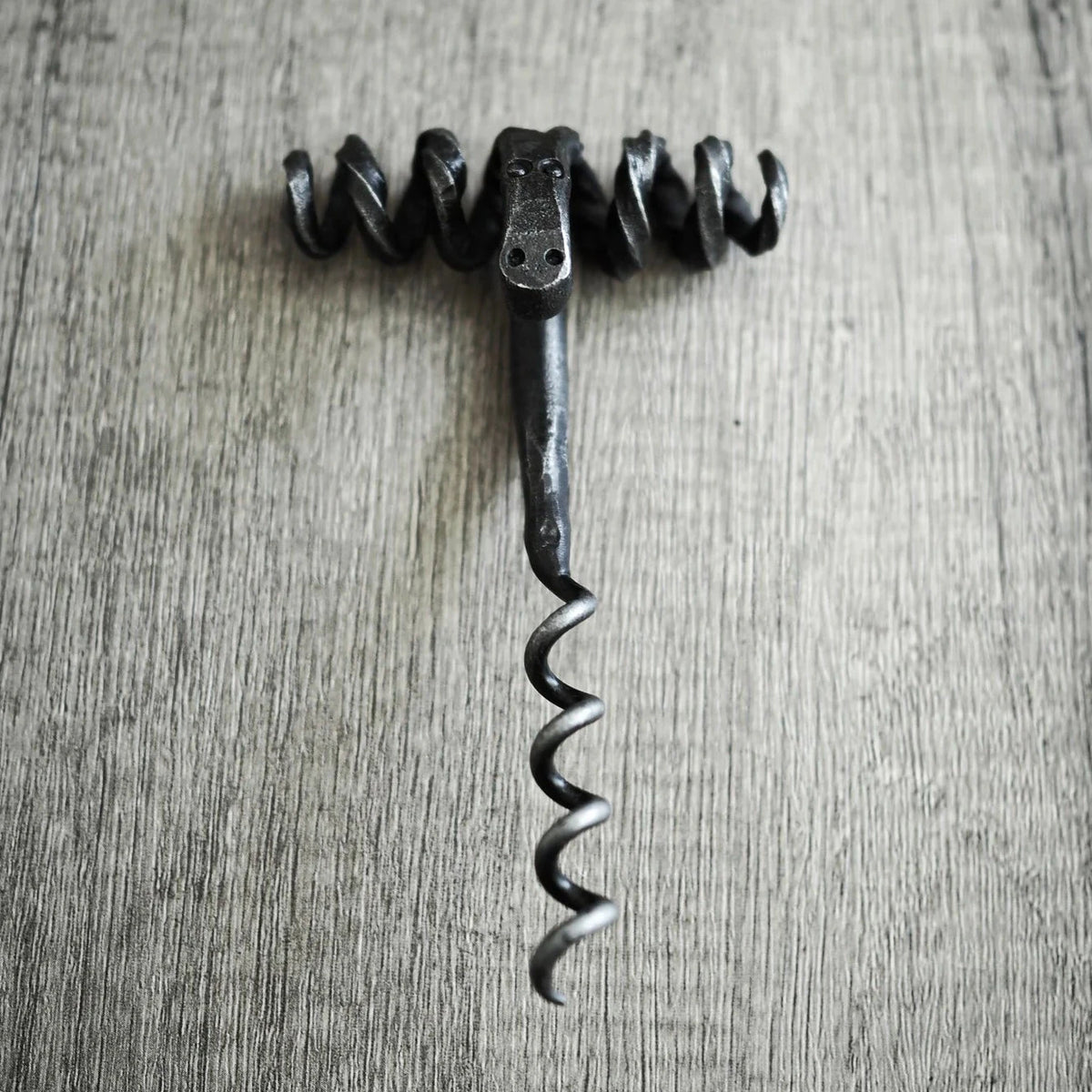 Bagge-skruv fotograferad ovanifrån på ljus träunderlag. Symmetrisk baggehuvud med framträdande böjda horn som sprider sig horisontellt. Öga och ansiktsfötryck synliga i metallhantverk. Spiral-skruv mekanismen arrangerad vertikalt nedan. Återvunnet järn i mörkfinish mot ljus bakgrund.