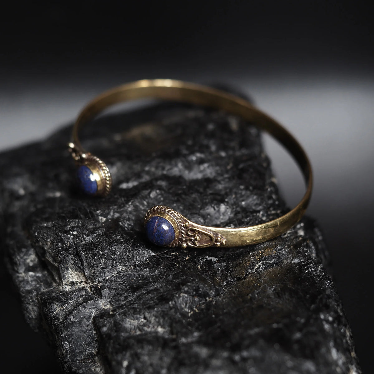 Mässingsarmband (doublearmband) med två infattade lapislazulistenar i ornamenterad mässingsfat. Öppen design med reparkeringdetaljer. Fotograferad på mörk stentextur.
