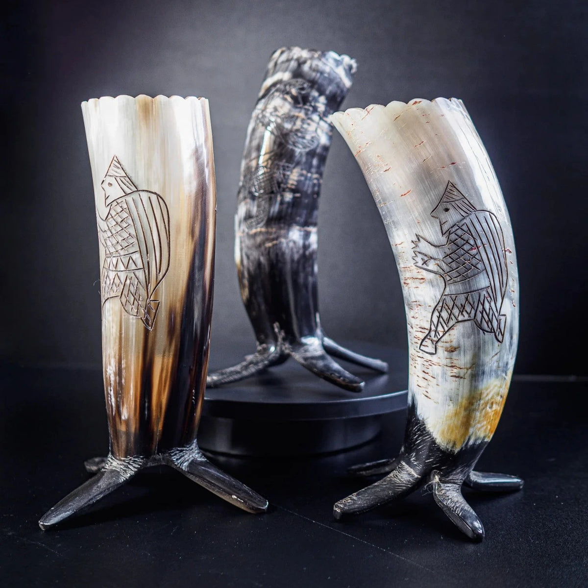 Tre organiska hornbägare med mörkbrunt patina, uppställda på metallstativ. Vänster och höger bägare har graverade vikingaskepp med geometriska mönster och skalliknande ornament som representerar segel. Mittbägaren visar naturlig hornstruktur med vågig kant.