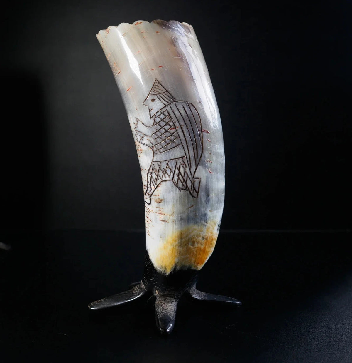 En organisk hornbägare visad i profil med mörk patina. Detaljer visar graverad vikingaskepp med geometriska sälmönster och komplext interlacing. Vågig kant synlig högst upp. Metallstativ vid basen.