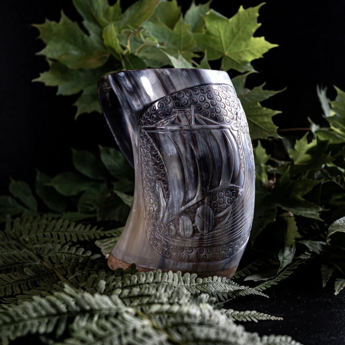Handsnidad hornmugg med vikingskepp-gravyr omgiven av ormbunks- och ekblad, visar intrikat traditionell utskärning av långskepp-design.