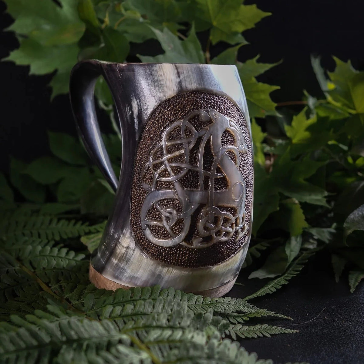 Två handsnidadehornnmuggar med Urnes-drakedesign bredvid varandra omgivna av ormbunksblad, visar traditionell vikingarnas drakkonst.