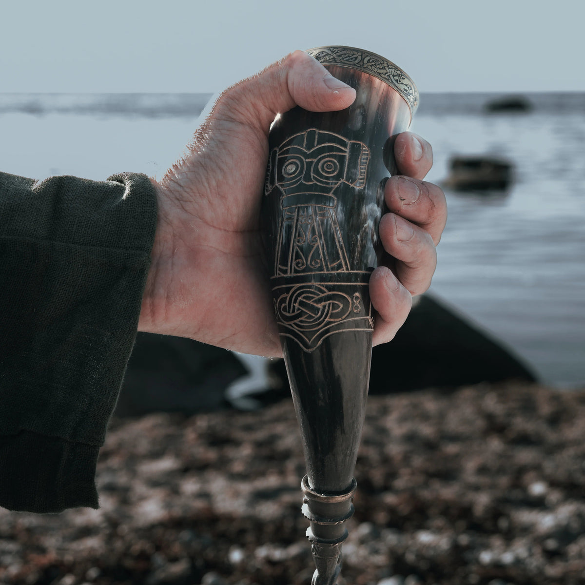 Dryckeshorn med korp och gravyr gjord av organiskt horn, cirka 250 ml, med mässingband och Viking-inspireade mönster, hållen i hand mot hav och klippor i bakgrunden