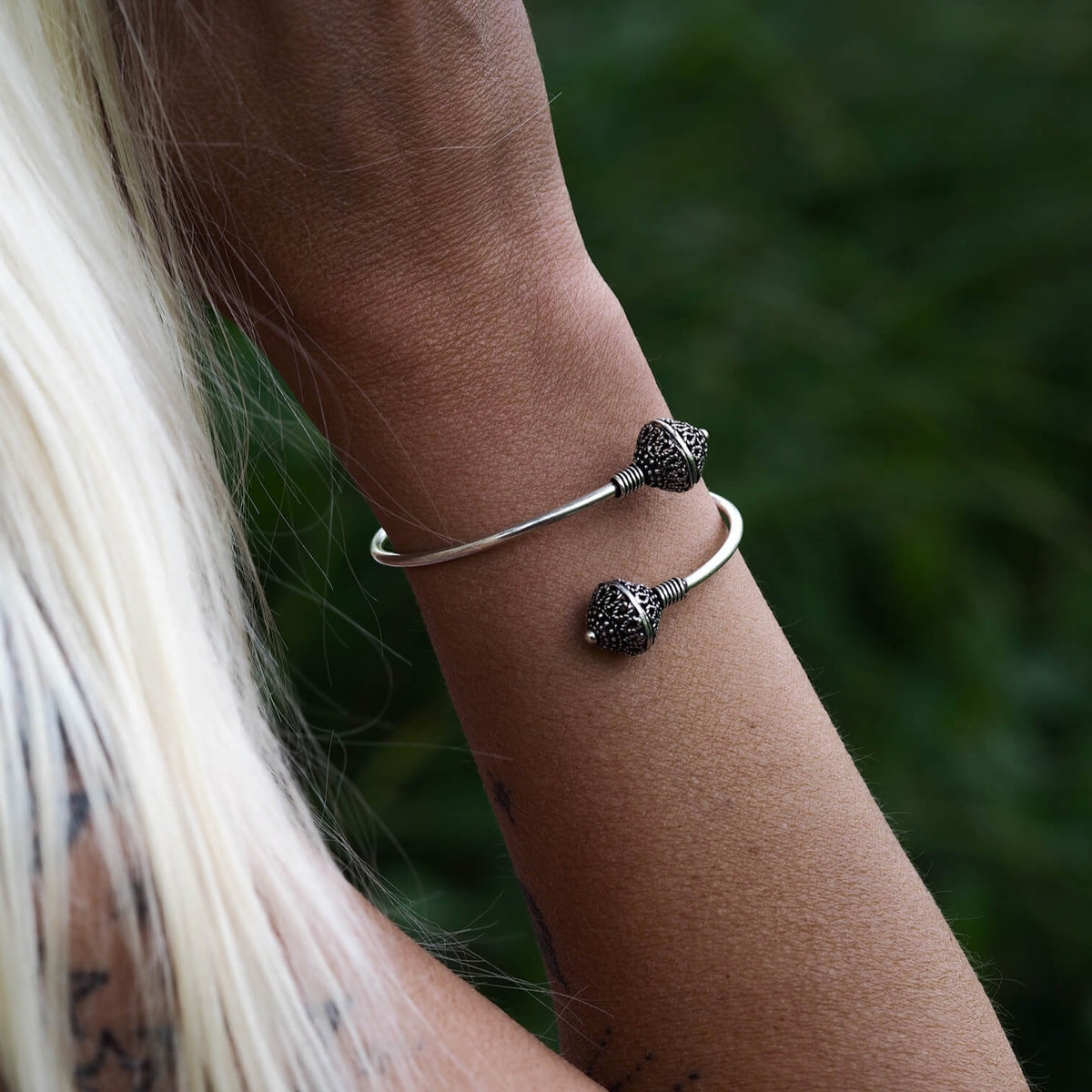 Silverpläterad mässingarmband med två pärlor på mörkbakgrund. Öppen armring med två graverade pärlor i mässing som visar vikingatida motiv. Polerad finish med varma guldtoner. Tråd-lindningar vid anslutningspunkterna demonstrerar traditionell metallteknik.