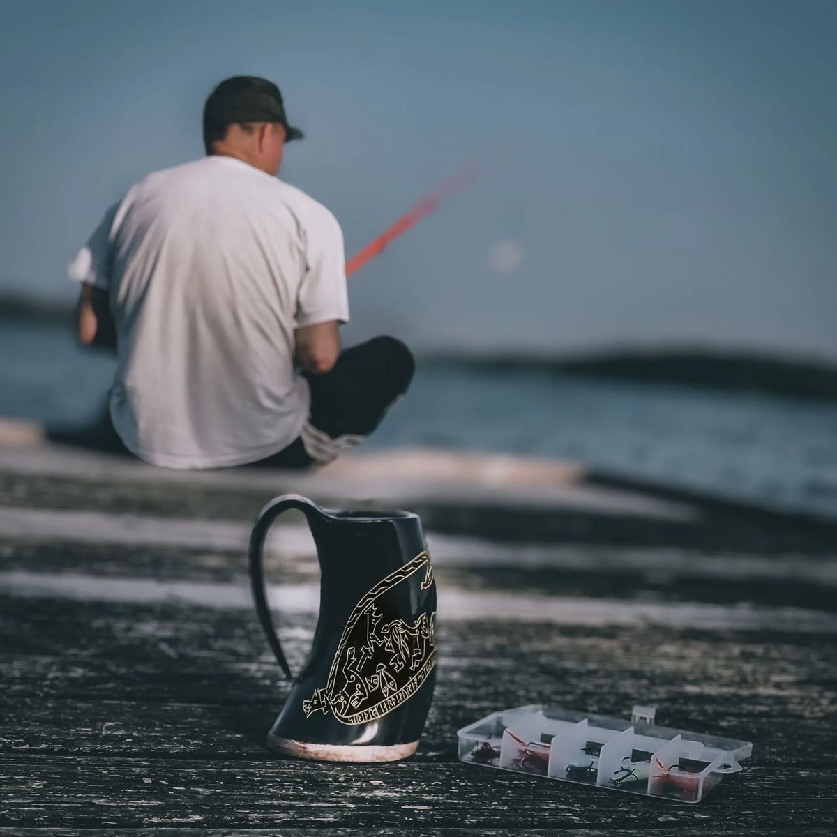 Två hornmuggar med Sigurdsagan-gravering på stranden. Naturligt hornmaterial med mörka graverade motiv. Mörkerivet hav och himmel skapar atmosfär av norsk sjöfartstradition.
