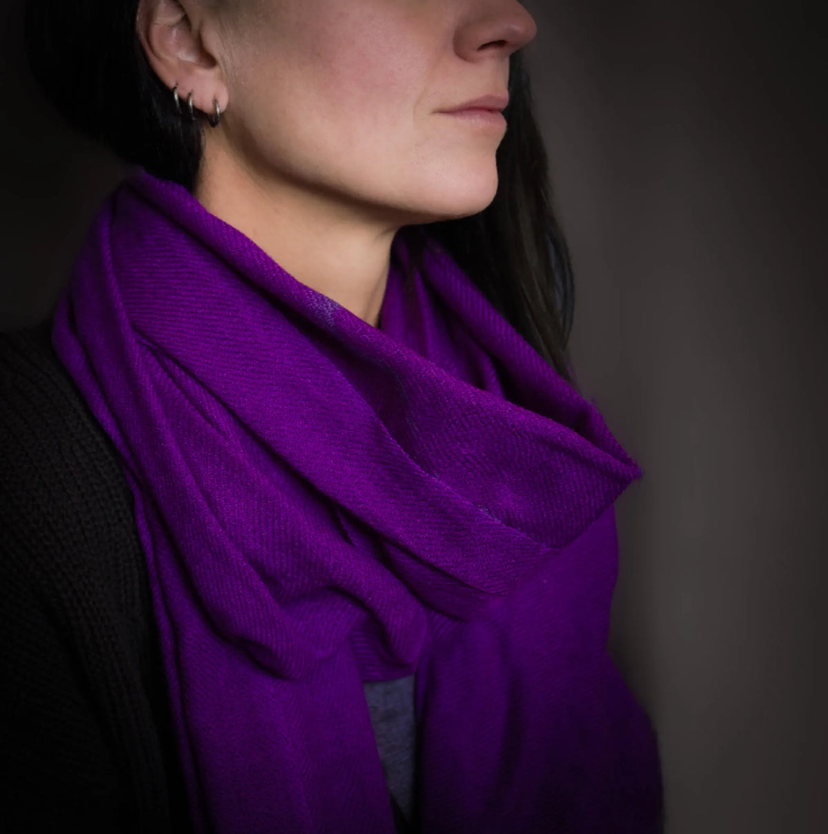 Lilla kashmirsjal bæres af model i profilvisning mod mørk baggrund, viser blødt materiale, elegant drapering og rig purpurfarve med dramatisk belysning.