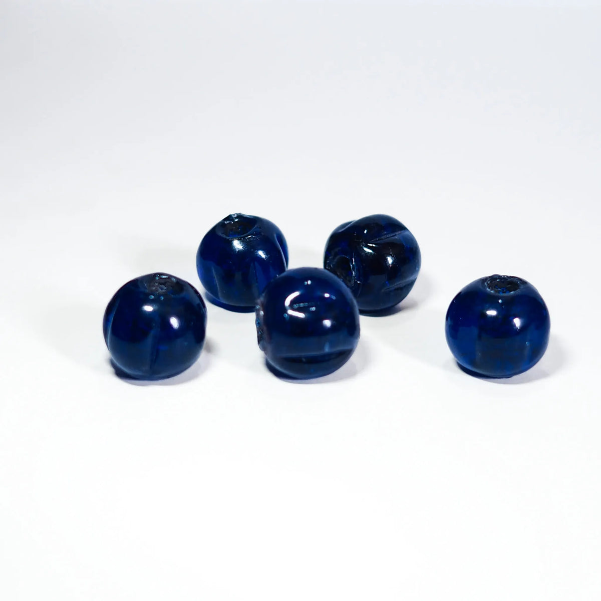 Blå melonpärlor i glas mot vit bakgrund. Fem pärlor visar mörkblå färg och naturlig variation. Central hål är synligt. Material: glas. Användning: pärlor för smycken och armband.