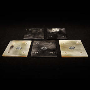 Fem kvadratiska hornbrickor för brickväv framlagda på vit bakgrund, med naturliga horn-färgningar från ljusbeige till mörkbrun, med inristade märken på varje bricka