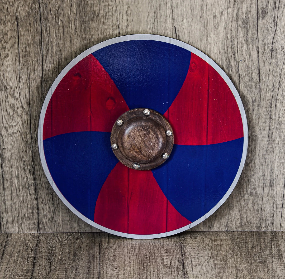 Vikingsköld för barn med röd och blå spiralmönster. En dynamisk spiral i rött och mörkblå sträcker sig från centrum. Brun träsköldbuckla med metallnitar i mitten. Vita kantremsor. Ligger på grå träyta.