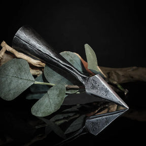 Smiderad järnspjuthållare bland torkade eukalyptusblad och växtmaterial, horisontell placering mot mörk bakgrund