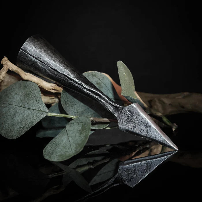 Smiderad järnspjuthållare bland torkade eukalyptusblad och växtmaterial, horisontell placering mot mörk bakgrund