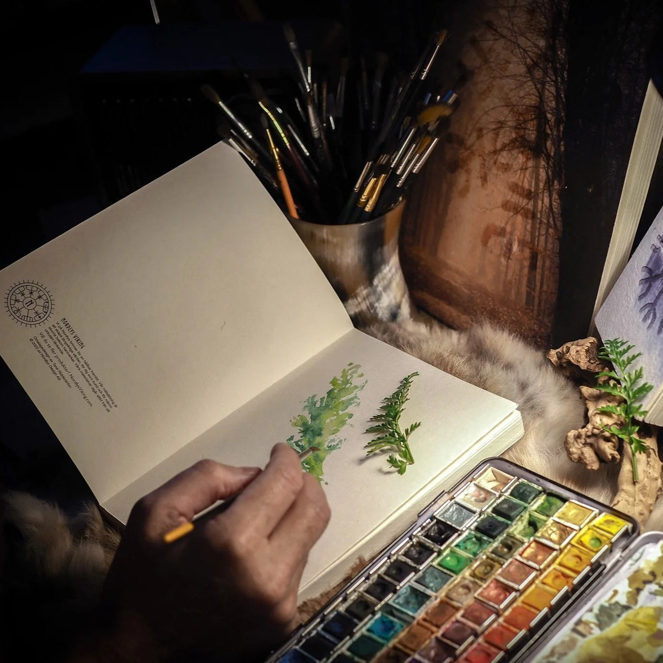 Målarbok A6 med Skyddsruna Aegishjalmur på omslaget. Bilden visar en öppen målarbok med blå-gröna växtillustrationer, målarpensel i hand, palett med vattenfärger och dekorativa växter runt omkring.