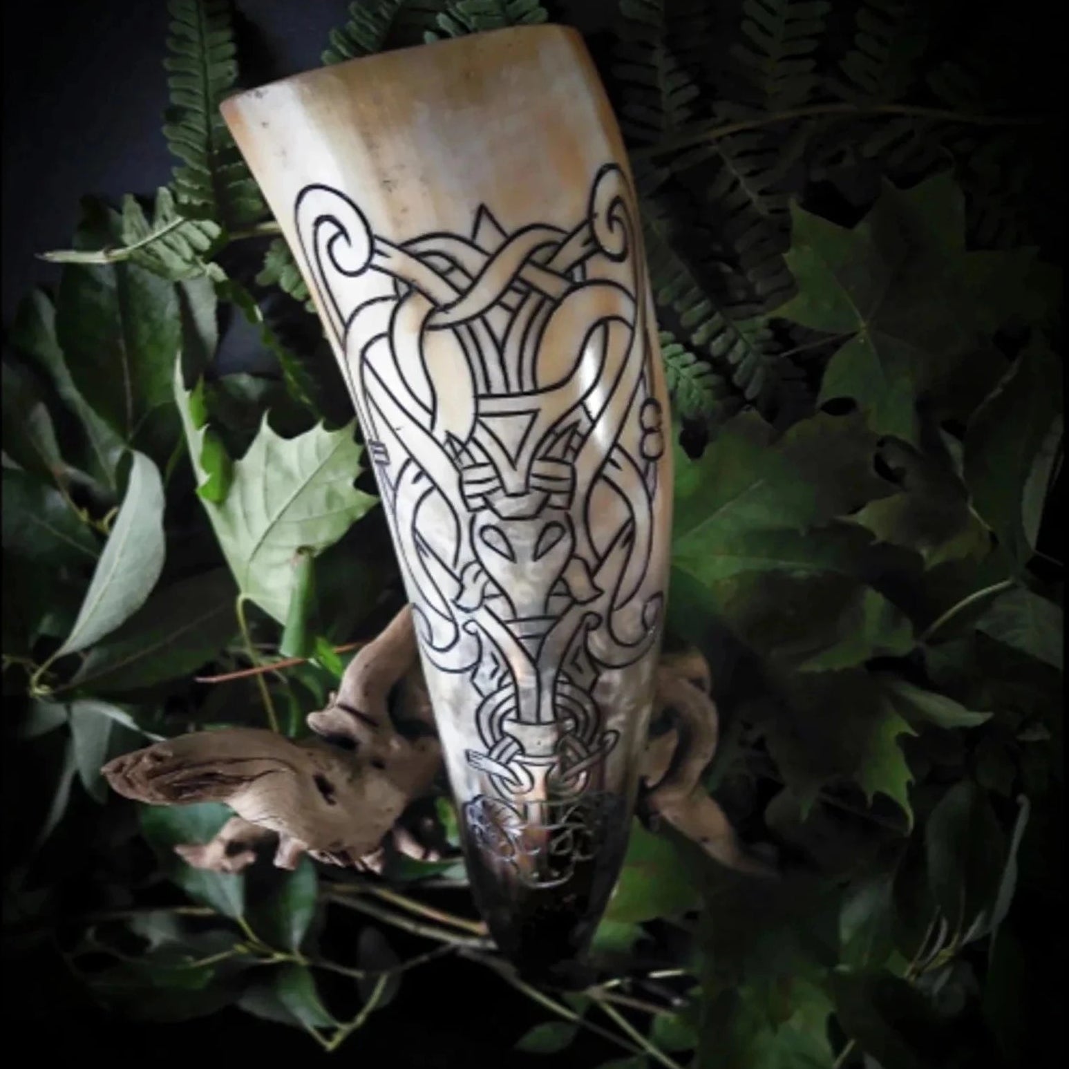 En organisk hornbägare med gräddvit och mörkbrunt patina, fotograferad bland gröna ormbunksblad. Gravering visar komplext norrskt knotwork och geometriska mönster som går runt hela hornets omkrets.