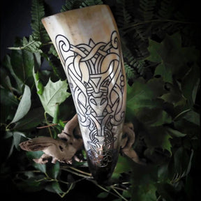 En organisk hornbägare med gräddvit och mörkbrunt patina, fotograferad bland gröna ormbunksblad. Gravering visar komplext norrskt knotwork och geometriska mönster som går runt hela hornets omkrets.