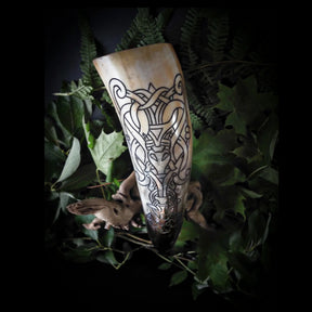 Naturligt bockhornfört med detaljerade gravererade nordiska knutmönster. Hornet visar naturliga bärnsten- och beigefärgningar med svarta graveringslinjer. Komplext keltiskt inspirerad design med symmetriska inflätningsmönster. Fotograferat vertikalt mot mörkbakgrund omgivet av grön ormbunkvegetation.