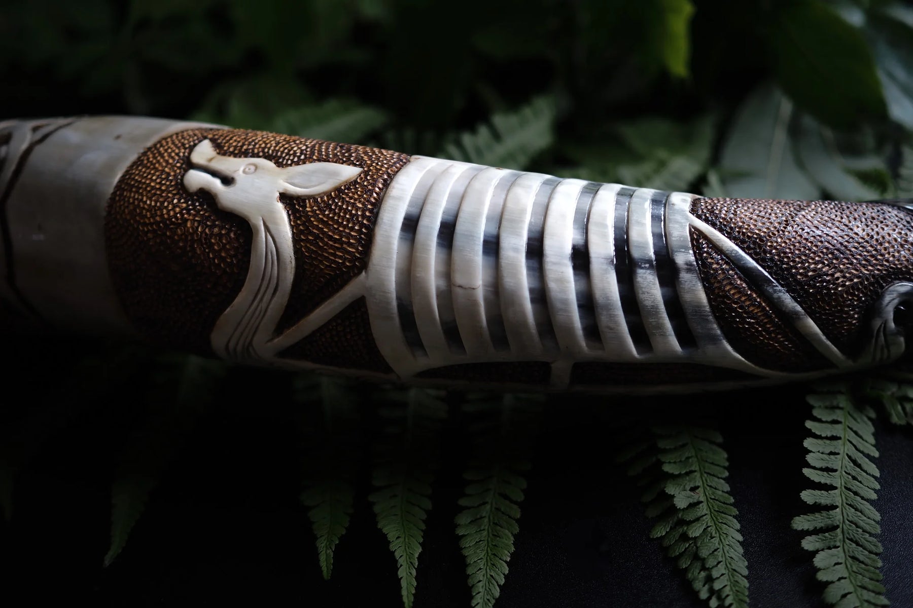 Närbild av organiskt dryckeshorn med minutiöst graverad vikingaskepp, vilande på grön växtlighet. Metallbanden och hornets naturliga struktur är i fokus.