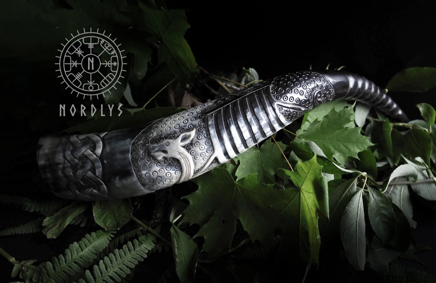 Dramatisk livsstilsfotografi av händer som håller dryckeshorn med vikingaskepp pyrografi mot grönska, visar både användning och dekorativ värde.