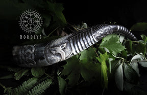 Dramatisk livsstilsfotografi av händer som håller dryckeshorn med vikingaskepp pyrografi mot grönska, visar både användning och dekorativ värde.