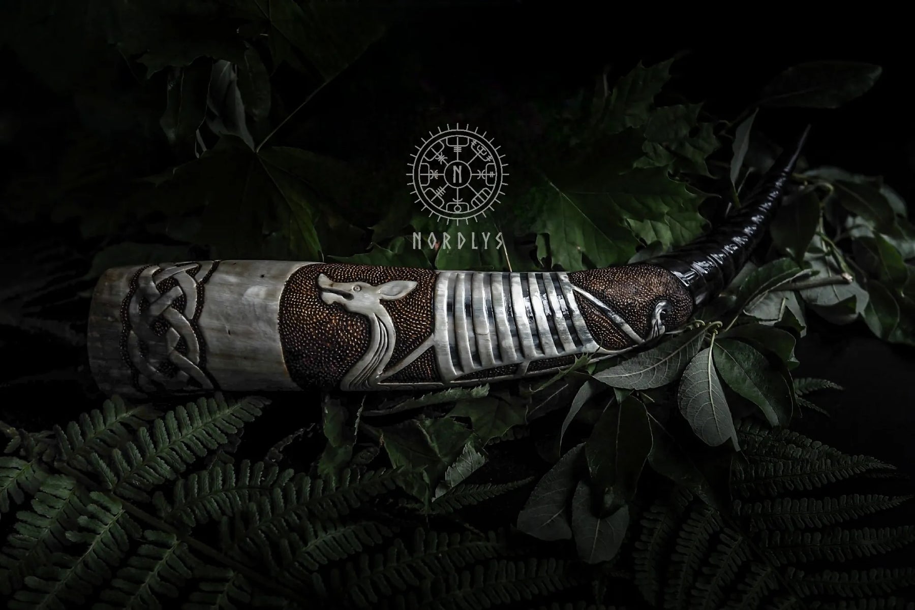 Djupt snidad dryckeshorn av organiskt horn med graverad vikingaskepp, mörkbrun läderband och metallfästen. Fotograferad mot naturlig bakgrund med gröna växter och Nordlys-logotyp.
