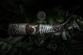 Djupt snidad dryckeshorn av organiskt horn med graverad vikingaskepp, mörkbrun läderband och metallfästen. Fotograferad mot naturlig bakgrund med gröna växter och Nordlys-logotyp.