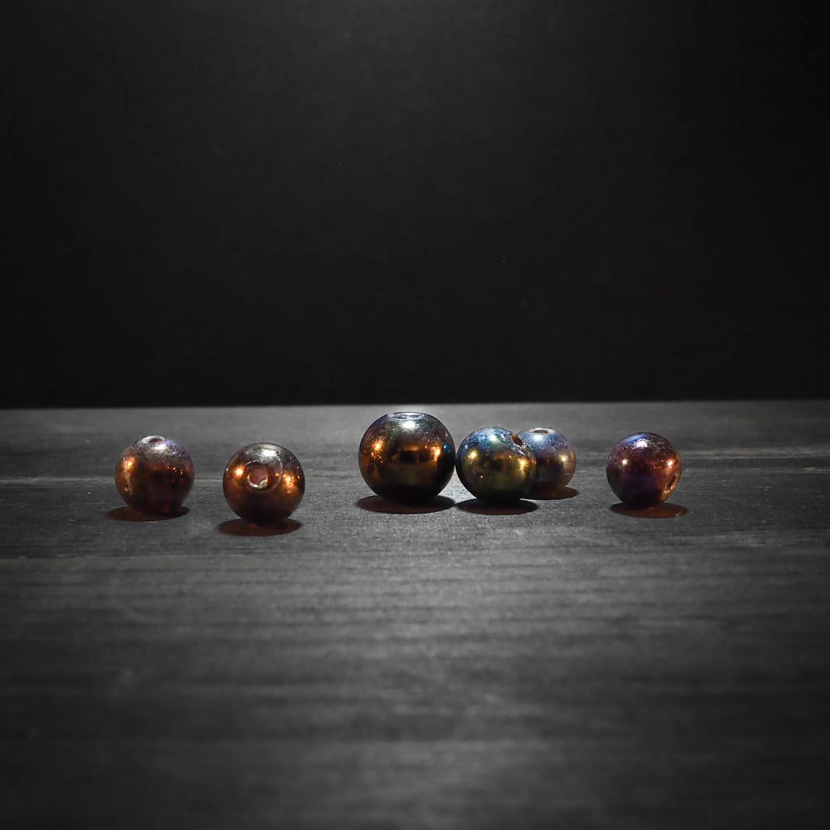 Sex glaspärlor med metallisk koppar-brunt glans, 7-10mm diameter, fotograferade på mörkgrå yta med dramatisk belysning