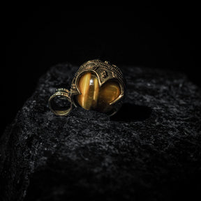 Mässinghänge gotlandskula med tigeröga på mörk stenig yta. Tigeroögestan med varma guldtoner är omgiven av en finmönstrad mässingsmontering med vikingiska ornamentala hörndetaljer och klassisk nordisk design.