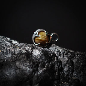 Gotländsk mässinghänge med tigeröga-sten på ljus stenbakgrund, visar stenens naturliga mönster och guldfärgad glans