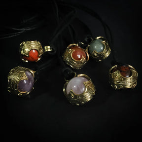 Sex Gotlandskula-hängen i mässing med varierade halvädla stenar på svart lädersnöre, visar olika färger av ametist, karnelsten och andra mineraler