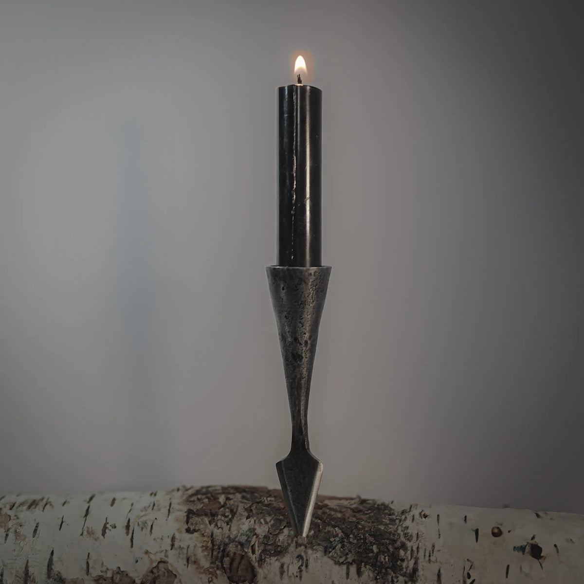 Gugnir spjut ljusstake av handsmide jarn presenterad i natural miljö med tent ljus och omgivande växter, detaljer visar forgjord finish och viking-inspirerad design