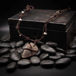 Stenpilspetshalsbandet presenterat på mörkträ och svarta stenar. Polerad beige-grå pilspets lindad med mörkt snöre med små benärlor längs läderband. Studiofotografi med dramatisk belysning som belyser stenens mineraliska textur och hantverk.