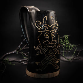 Mörk ölmugg i horn med graverat Odensmask-motiv i guld, mot svart bakgrund med kvistar och lav