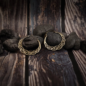 Två mässingörhängen med keltisk flätad knutdesign i guld/messingstyrka. Detaljerade sammanvävda knute visar på träunderlag med stenar.