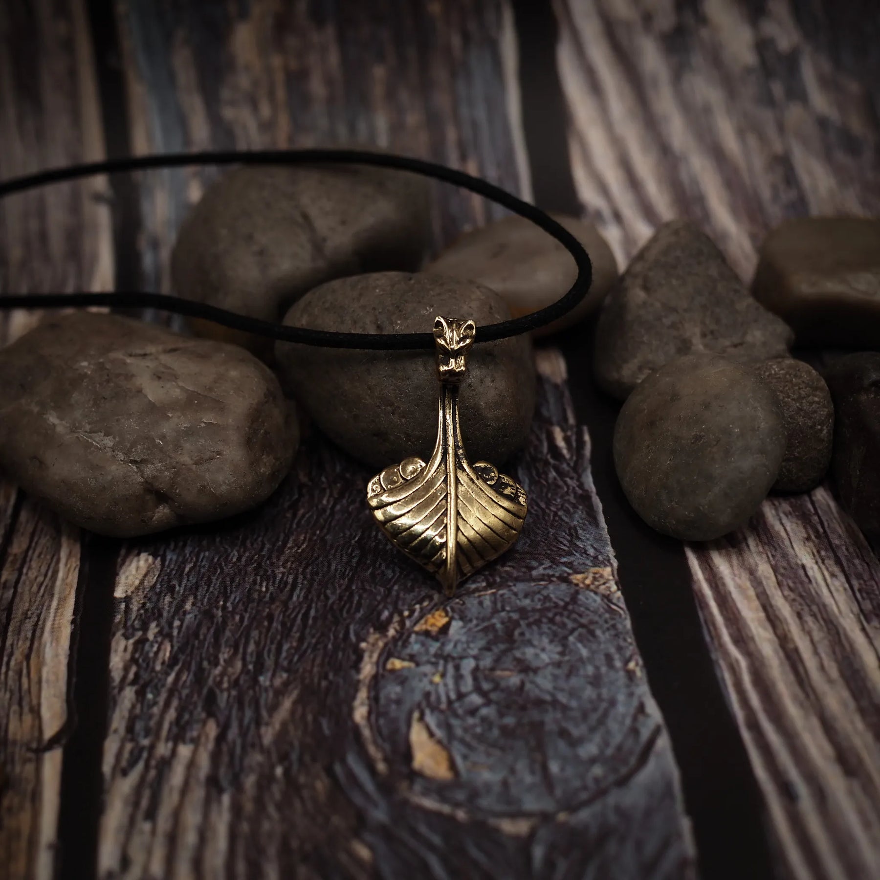 Vikingaskepp-hänge på svart lädersnöre placerat på grå stenar och träyta. Brons med varm patina och drakehuvud synligt.