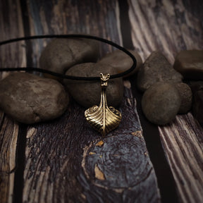 Vikingaskepp-hänge på svart lädersnöre placerat på grå stenar och träyta. Brons med varm patina och drakehuvud synligt.