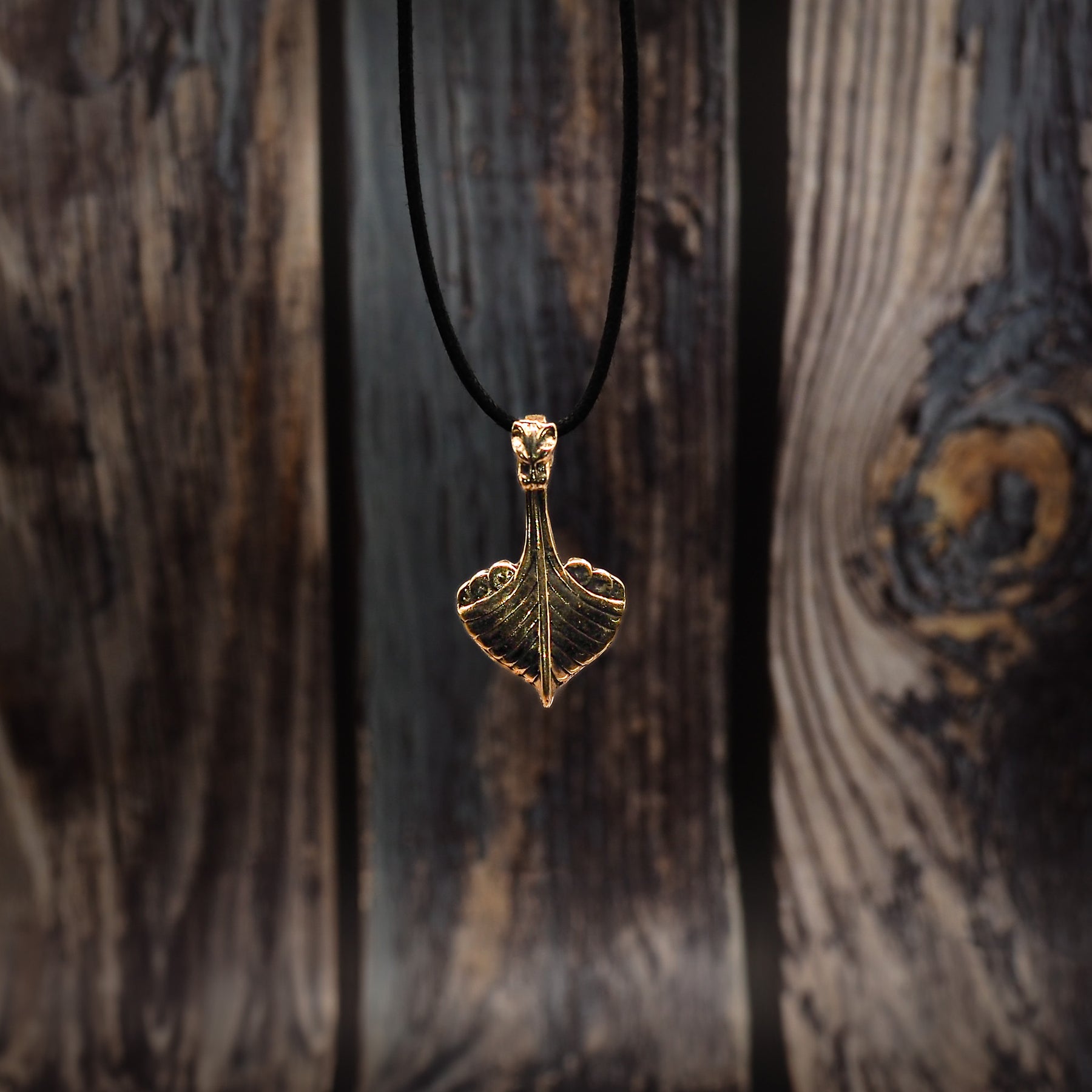 Vikingaskepp-hänge i brons i frontvy mot träbakgrund med runor. Tydlig drakehuvud och skeppskrov i metallguss.