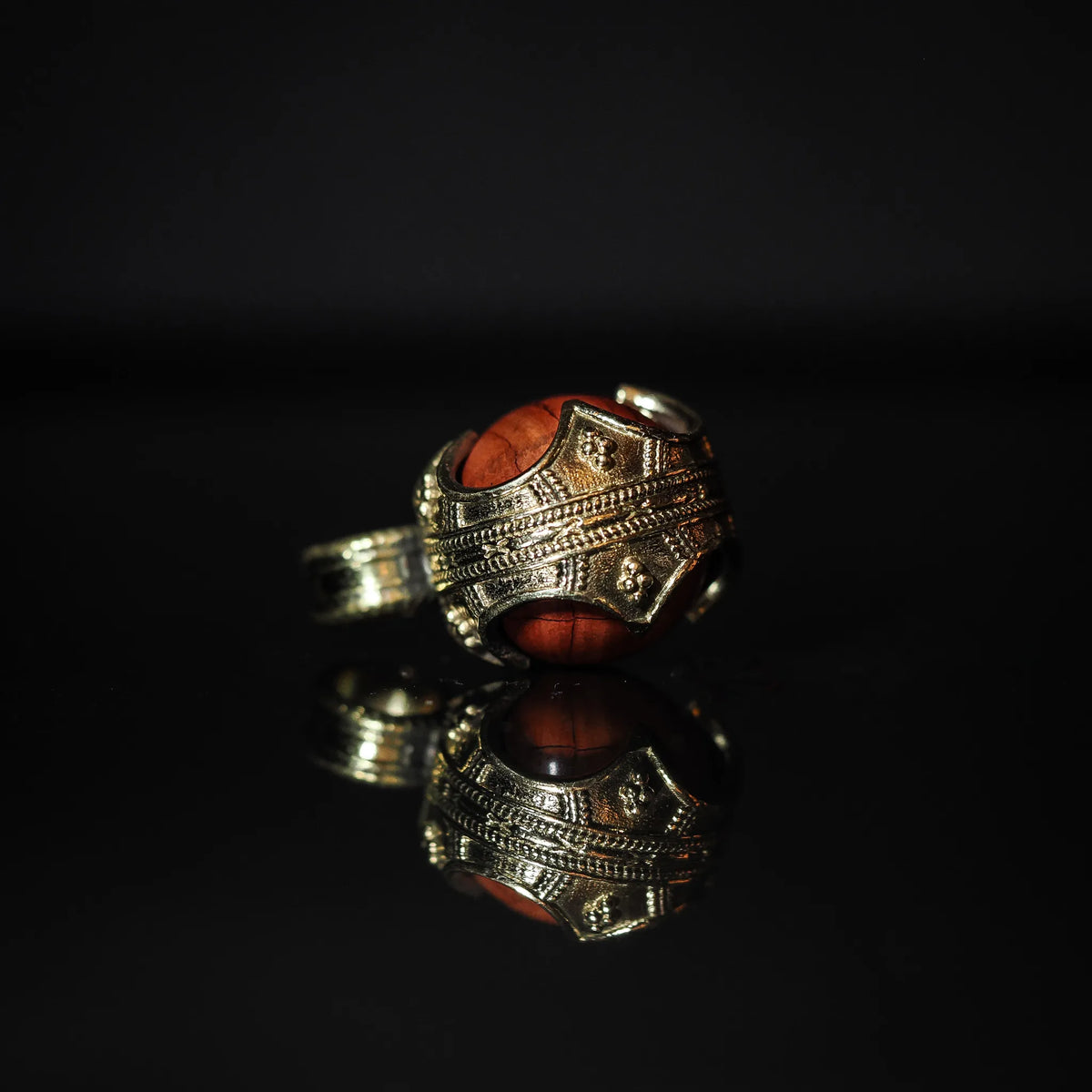 Närbild av mässingshänge med röd jaspis-sten. Ornerad brassmålade metall med detaljerade linjemönster. Profilvyn visar glansig röd jaspis i klassisk vikingahängeskapsyl.