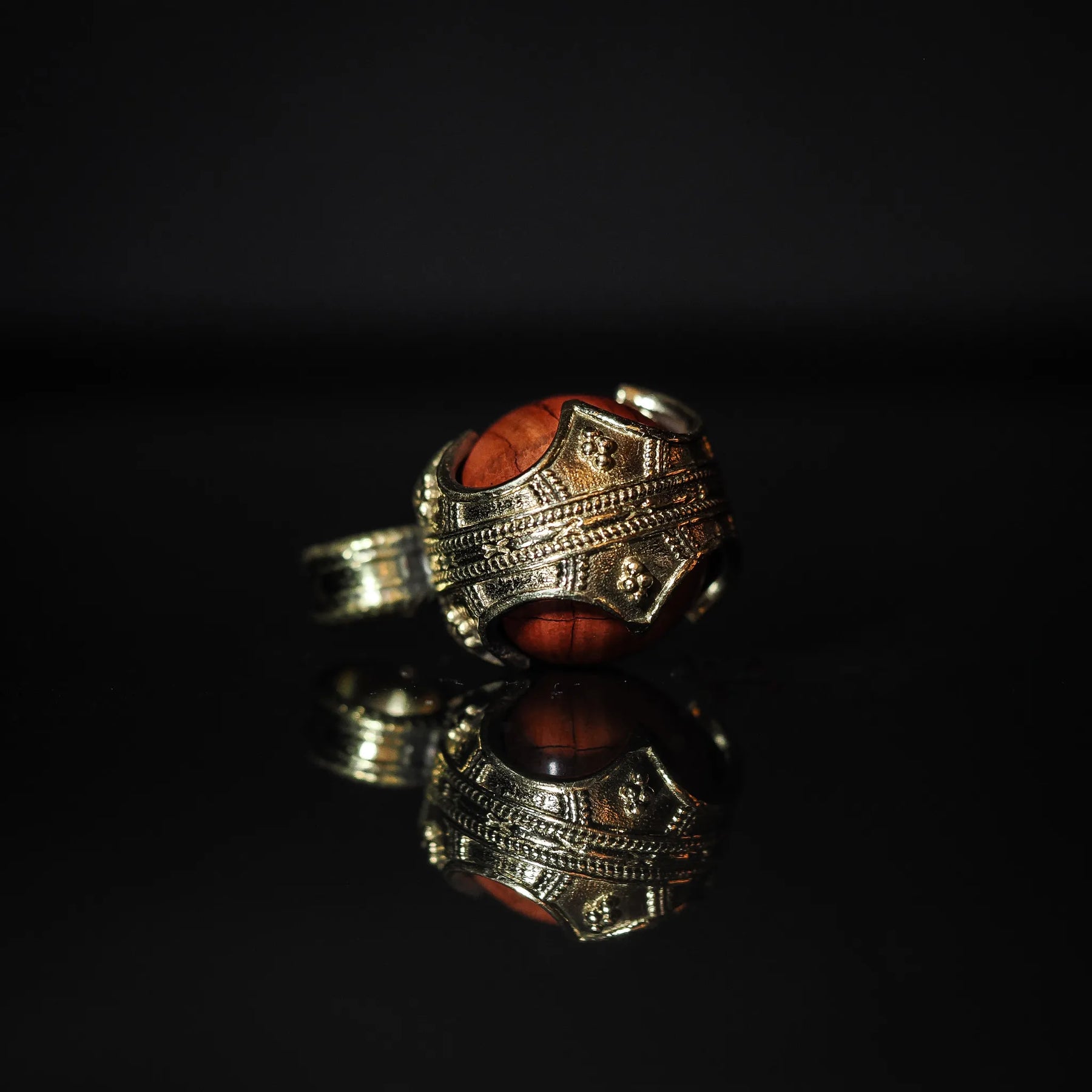 Närbild av mässingshänge med röd jaspis-sten. Ornerad brassmålade metall med detaljerade linjemönster. Profilvyn visar glansig röd jaspis i klassisk vikingahängeskapsyl.