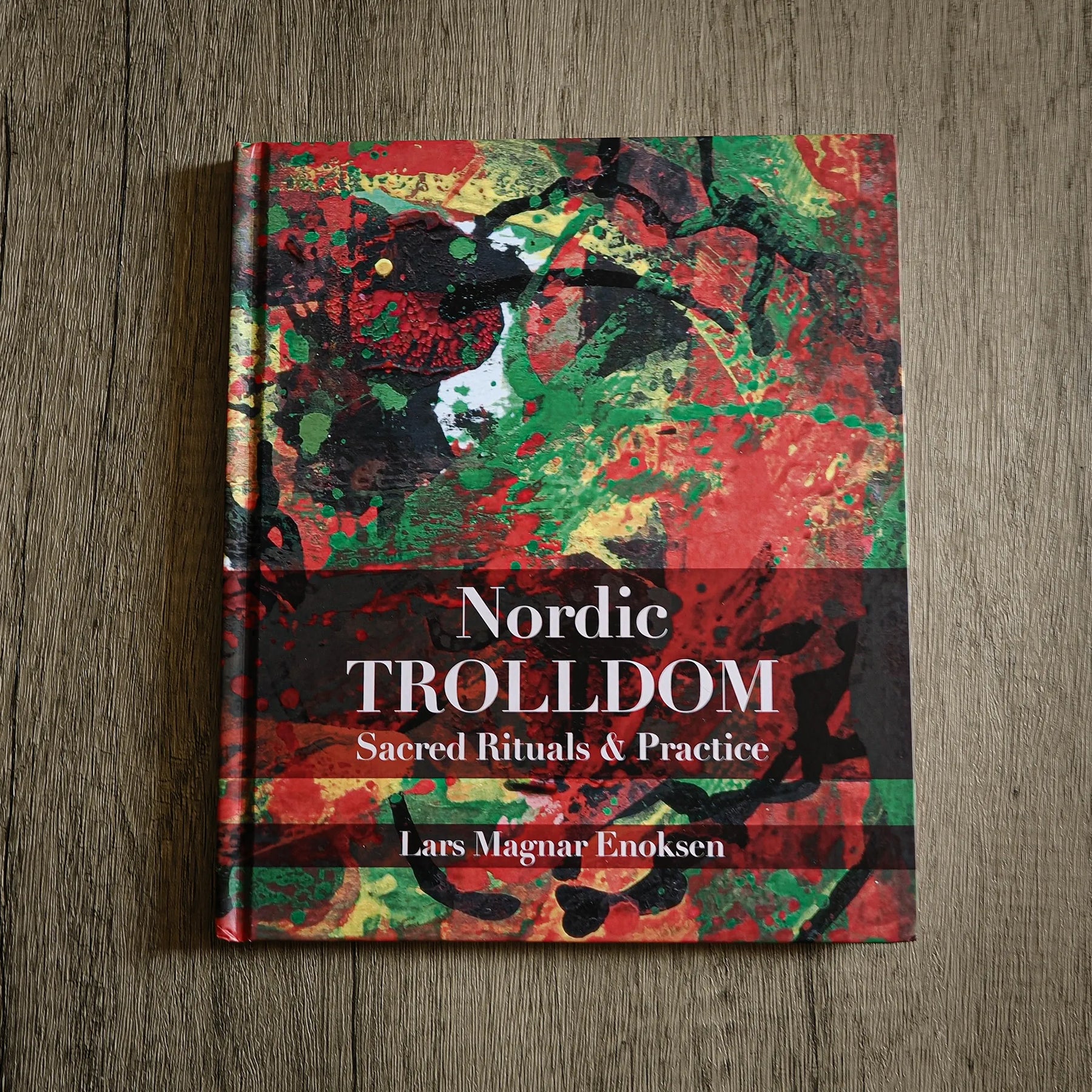 Boken Nordic Trolldom av Lars Magnar Enoksen, hårt omslag med abstrakt rödgrönt konstverk, på träyta