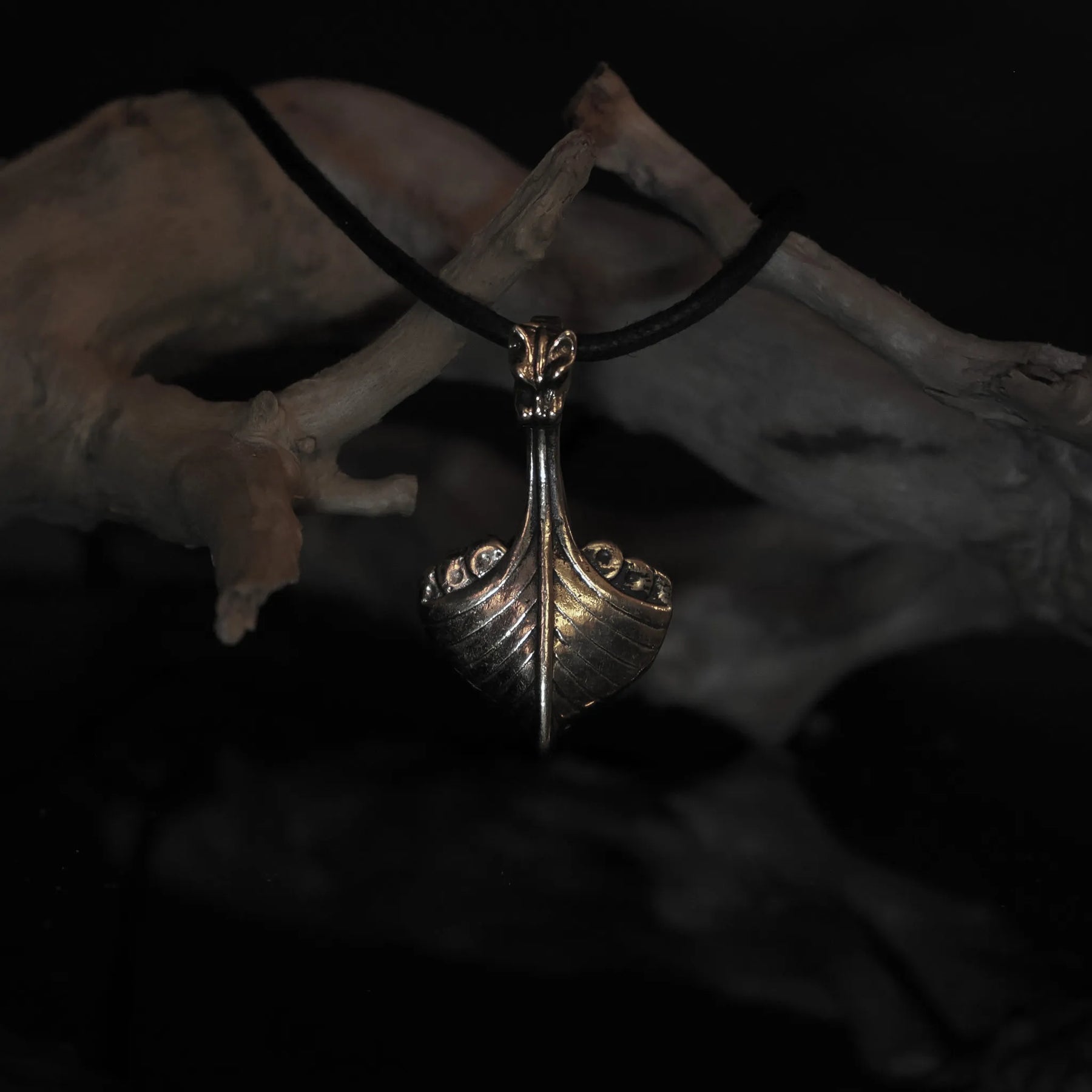 Vikingaskepp-hänge i brons från vinkelperspektiv. Väl utformad drakehuvudsprö och hölskonstruktion. Svart läderband.