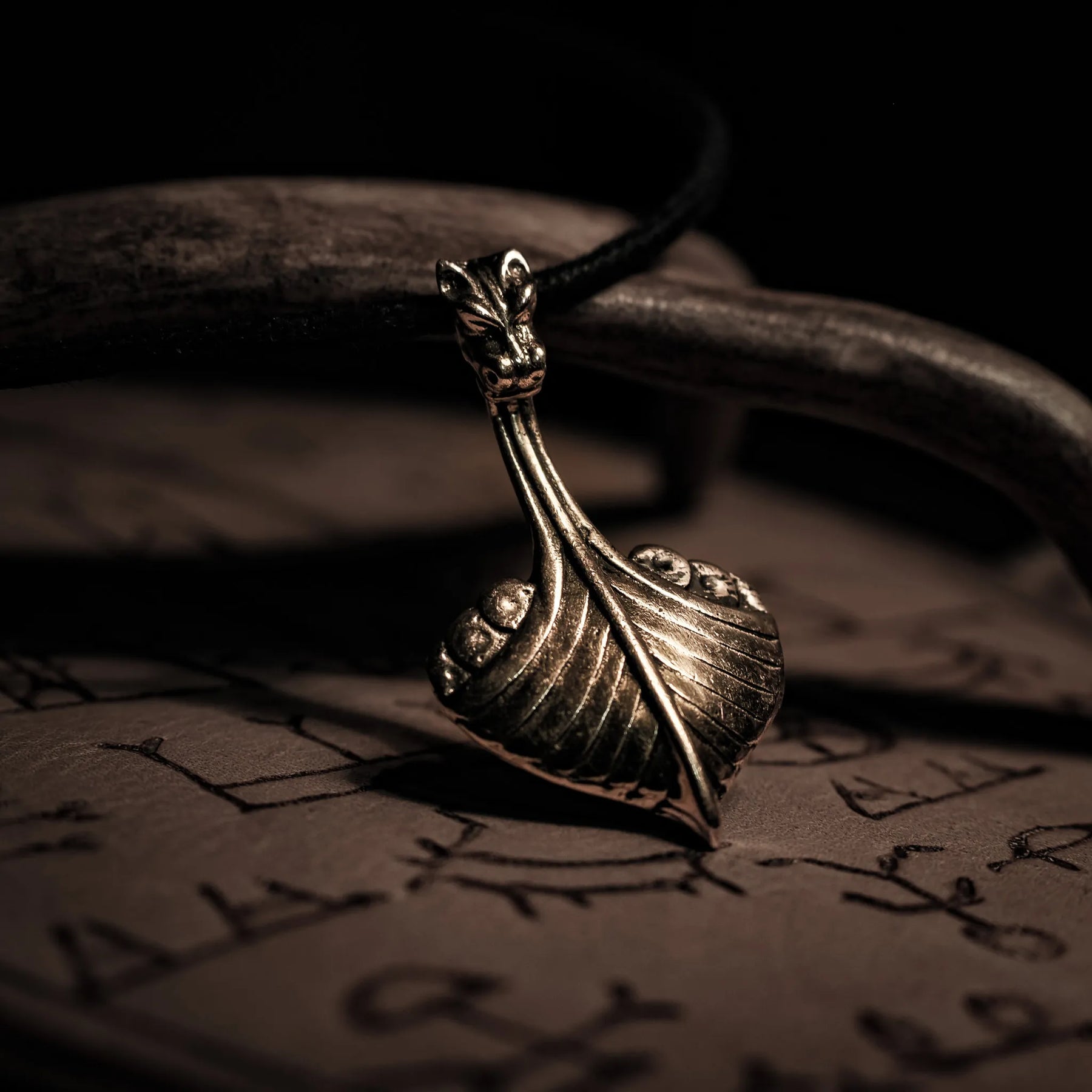 Bronshänge i form av vikingaskepp med drakehuvud vid förskepp. Hängande från svart lädersnöre. Detaljerad metallarbete med varm glans.