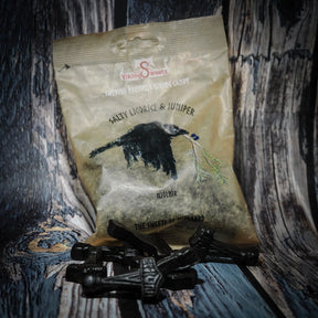 Svarta Mjölnir-formade godisbitar framför en pastellgul förpackning med en svart råkorre och grön enriskvista. Vikinggodis i saltlakrits- och enrismaK presenterad på trä.
