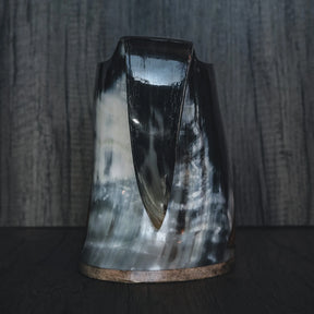 Frontvy av handsnidad hornmugg med konisk form och naturlig hornfärg. Visar den karakteristiska spetsiga basen och hornets naturliga kornstruktur. Trädäband vid kanten. Tjockt, organiskt horn utan gravering.