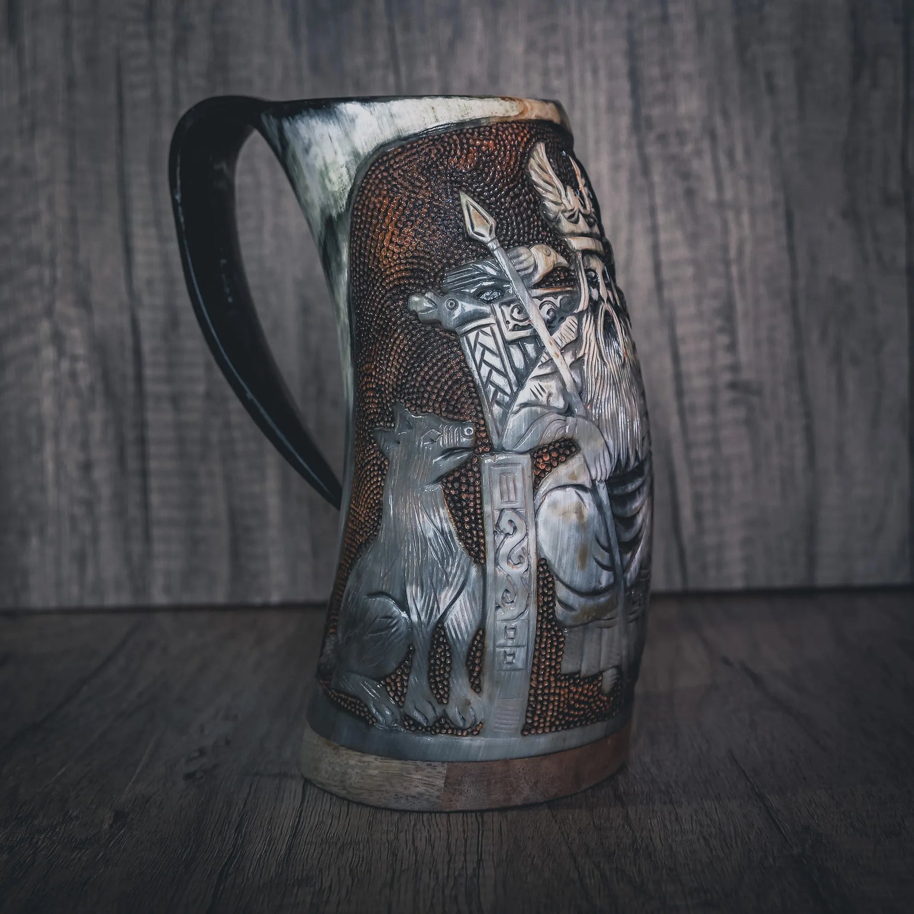 Handsnidad hornmugg fotograferad från sidan med fullständig Odin-figur synlig. Stående krigar i full detalj hållande stav eller spjut med elaborerad klädsel och hjälm. Omfattande mönsterarbete och dekorativa element omger huvudfiguren. Naturligt horn med utmärkt sniddjup. Kopparband vid basen. Dramatisk belysning framhäver den höga reliefsnidningen.