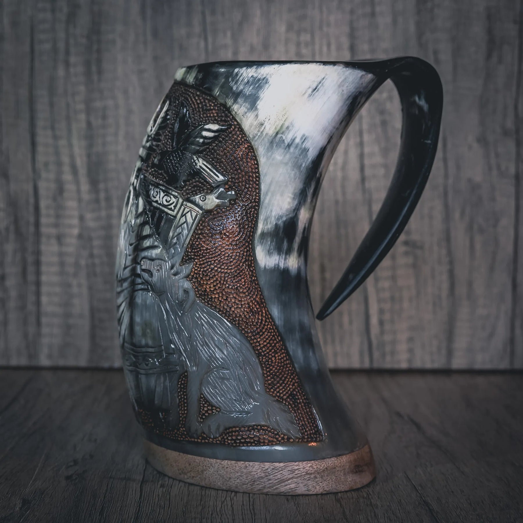 Handsnidad hornmugg visad från sidan med detaljerad reliefsnidning av Odin-ansikt. Bred skägg och intrikat mönsterarbete på ytan. Naturligt horn med varierande färg. Mörkad handtag och kopparband vid basen. Studiolysning belyser sniddetaljerna tydligt.