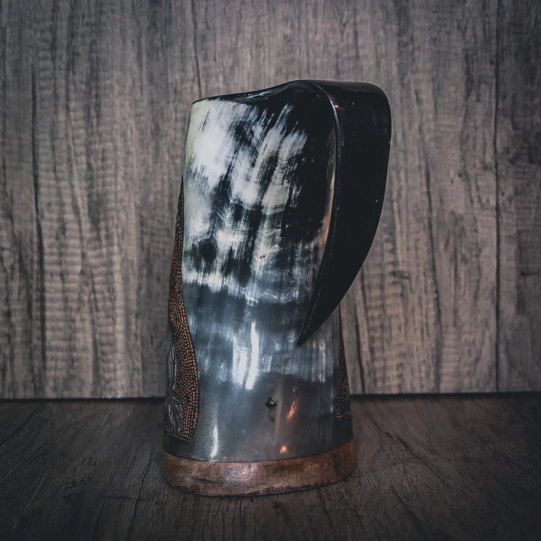Handsnidad hornmugg fotograferad från sidovinkeln. Visar naturliga hornådror och färgvariationer. Mörkad handtag och kopparband vid basen. Subtil reliefsnidning synlig på ytan. Konisk form med organisk hornstruktur framhävd genom professionell belysning.